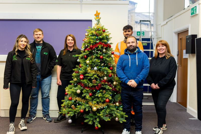 A group of BAM employees standing together in front of a decorated Christmas tree at Happy Days during a volunteering day. They are smiling and dressed casually, with a festive atmosphere in the background.