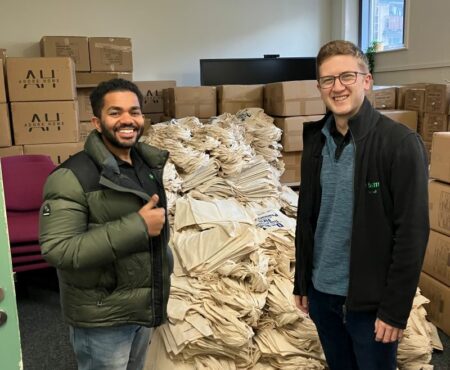Two men from BAM volunteering at VAC in Halifax, standing in front of a stack of boxes and tote bags. They are wearing casual clothing and smiling, with the boxes and bags likely containing supplies or donations.