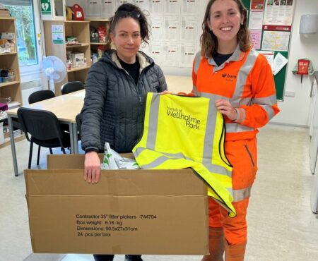 A BAM team member handing over high-vis jackets to a representative of the Wellholme Friends community group. Both individuals are smiling, they are at the project site. The jackets are bright yellow, symbolising safety.