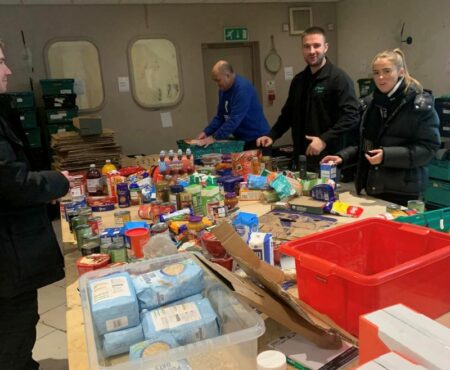 Volunteers gathered around a table, boxing up food items to support Focus for Hope in Brighouse. The table is filled with supplies like canned goods, dry foods, and packaging materials. The volunteers are actively engaged, working together in a spirit of community.