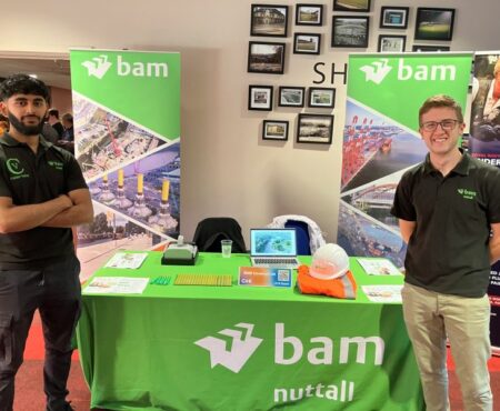 Two BAM staff members standing beside a display stall at an event. They are wearing branded clothing and smiling, with the stall showcasing informational materials, banners, and promotional items related to BAM’s work.