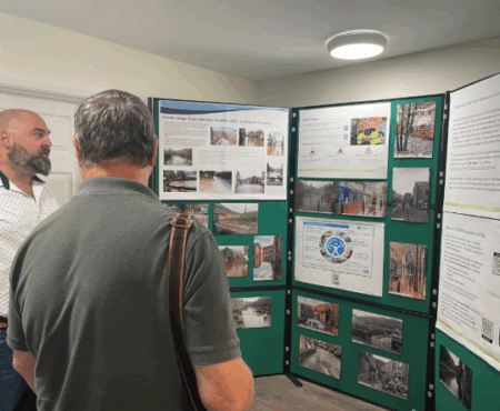 Two men standing and examining flood information boards. The boards display maps, diagrams, and text about local flood risks and safety measures. The men appear engaged and in discussion.