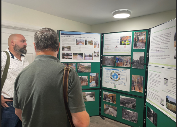 Two men standing and examining flood information boards. The boards display maps, diagrams, and text about local flood risks and safety measures. The men appear engaged and in discussion.